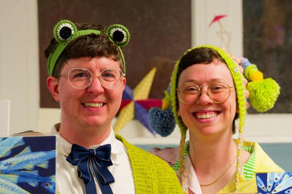 Man on left with frog eye handband and blue and green sleeves with woman to the right wearing a puffy headdress of similar construction,  both wearing glasses.  A spikey shape in primary colors rests on a wall with brown rectangles surrounded by white