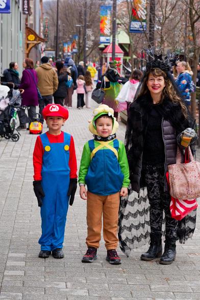 One boy wears a Mario costume and the boy to his right looks like Luigi and a woman to the right of him is wearing dark clothes and is holding a lot of bags with what looks like a substantial crowd behind them