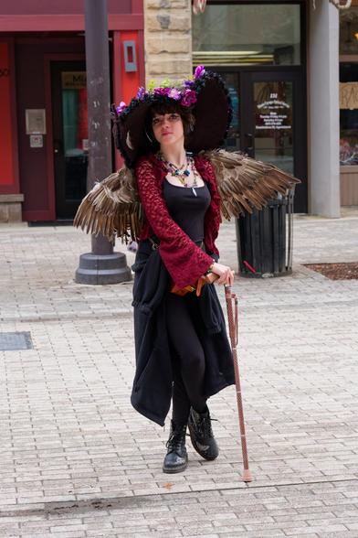 Fairy woman with big hat, feathered wings and a cane and dark dress.  she's standing with one toe pointing at the ground on a brick surface with storefronts and a trash can behind