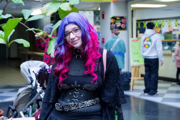 Woman with hair that is on the blue side of purple at the top with a gradient to the red side of purple in spirally drills wearing a black corseted dress with little metal hearts in front.  Behind her something like an umbrella tree and a vinyl floor with people in the background lined up for a restaurant