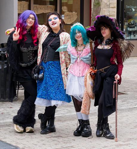 Four young women who can be seen elsewhere in the photo gallery pose together including:

goth with purple gradiant hair, face painted person with black blouse and medium blue skirt, pink white and blue fairy and dark fairy with crinkled witch hat