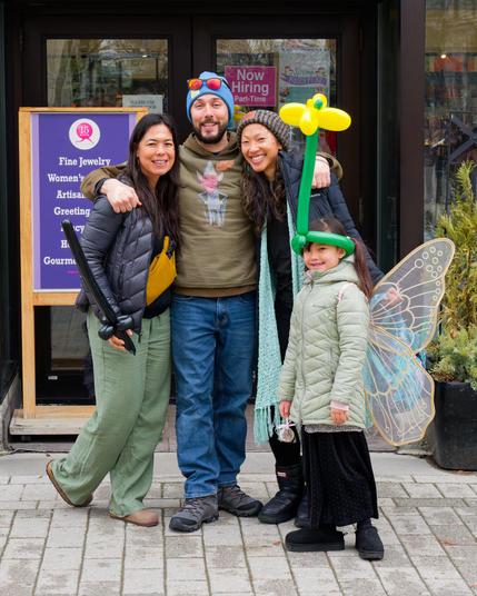 A man with a blue wool hat, brown shirt, jeans and bead has his arms around two dark-haired woman's shoulders, everyone smiling,  with a small girl wearing a puffer coat, butterfly wings and something made from balloons