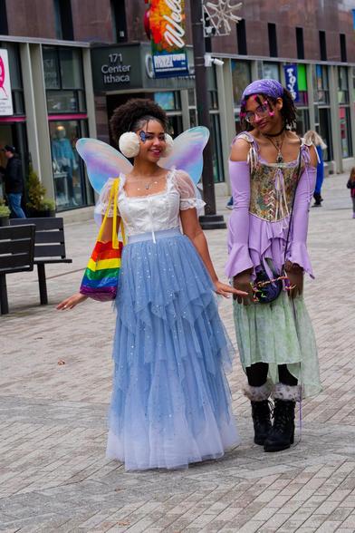 Two dark skinned woman dressed as fairies: on the left with a tiered blue skirt, white blouse, iridescent wings and rainbow bag and on the right purple blouse with detatched sleeves, purple headscatf, paisley corset and fur-trimmed boots.