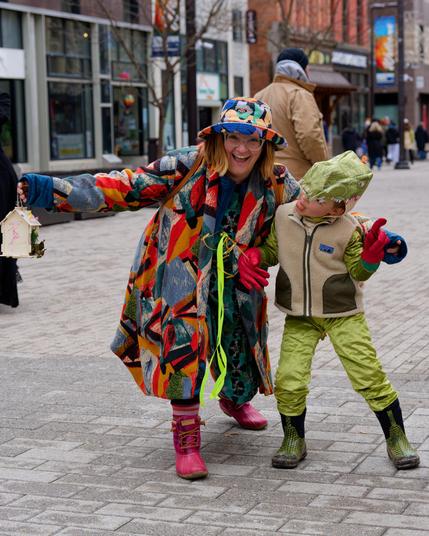 On the left a woman in a raincoat with many clashing primary colors and pink boots and a hat like her coat and red hair and big glasses and a big smile.  On the right a boy in green boots, green jacket, green sleeves, green hood,  all waterproof as well as red gloves