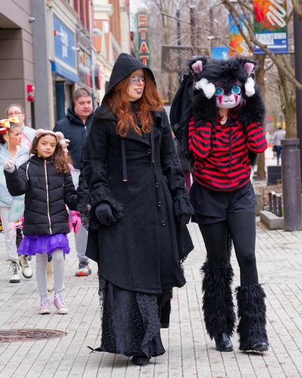 on the far left are some storefronts and a group of childen but the main event is a pair of woman,  to the left wearing a dark cloak and red hair and face paint and looking to the right and to the right as a stylized cat with red and black striped shirt, furry leggings and bat wings