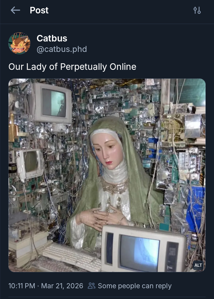 A screenshot of a social media post features a caption and an image depicting a religious statue surrounded by technology. The username displayed is "Catbus" with the handle "[@]catbus.phd," accompanied by a profile picture of a cartoon dragon. The caption reads "Our Lady of Perpetually Online" in white text against a dark blue background. The main image shows a statue of a figure in a white robe and green veil, standing with hands clasped in a prayerful gesture. This figure is surrounded by a dense, chaotic wall of electronic components, including green circuit boards, tangled wires, and beige CRT computer monitors. In the foreground, a large beige monitor and a keyboard are visible, while the post's timestamp indicates "Mar 21, 2026" at the bottom.