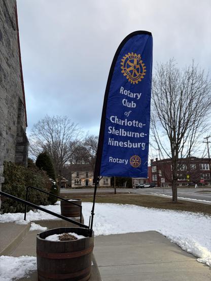 A flag for the Rotary Club of Charlotte-Shelburne-Hinesburg, featuring the Rotary emblem, stands near a stone building. It's winter, with snow on the ground and leafless trees in the background. A wooden barrel planter is visible,