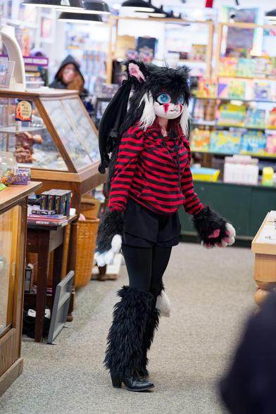 Woman wears black high heeled boots, furry leggings, black leggings, black skirt, furry cat gloves, red and black striped shirt and cat mask with bat wings behind her with a blurred out woman wearing a cloak waiting for her chance at the runway