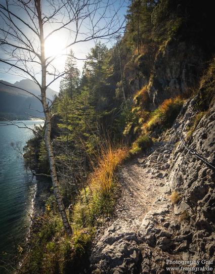 A breathtaking lakeside trail scene unfolds in this image, capturing the serene beauty of a rugged coastal path. The foreground features a narrow, rocky trail that winds along the edge of a steep cliff. The trail, bordered by patches of grass and shrubs, invites exploration and promises stunning views.

To the left of the trail, a calm lake stretches out, its tranquil waters reflecting the sunlight. The lake is nestled amidst the cliffs and trees, adding a sense of peaceful solitude to the scen…