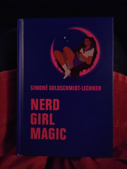 A photograph of the hardcover edition of Nerd Girl Magic by Simoné Goldschmidt-Lechner. The book is dark blue with pink letters and a magical girl reminiscent of Sailor Moon in the top half. 