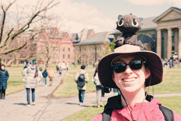 Woman with a boonie hat and dark glasses, red jacket and backpack straps has a brown dragon plush on her hat with big eyes