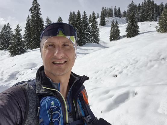 A vibrant and invigorating winter scene unfolds in this image, capturing a moment of adventure and joy in a snowy alpine landscape. The foreground features a hiker, smiling warmly at the camera, ready to embrace the beauty of the winter wilderness. The hiker is equipped with a backpack and dressed in outdoor gear, including a headband and sunglasses, prepared for a day of exploration.

The background showcases a stunning snowy slope, blanketed in pristine white snow that glistens under the brig…