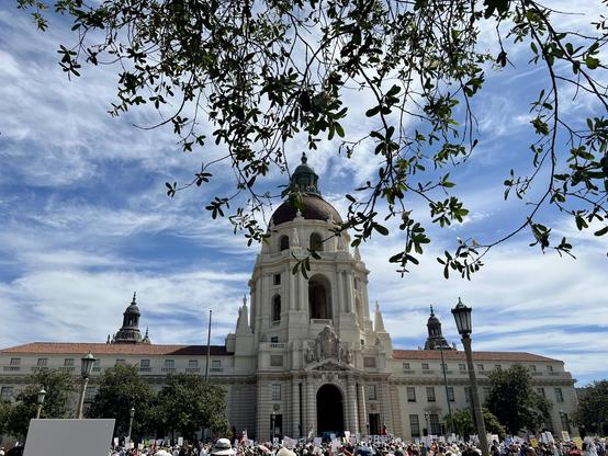 At NoKings protest, in front of Pasadena City Hall with Judy Chu speaking. The beautiful Mediterranean-style City Hall building is flanked by wispy high clouds in a blue sky