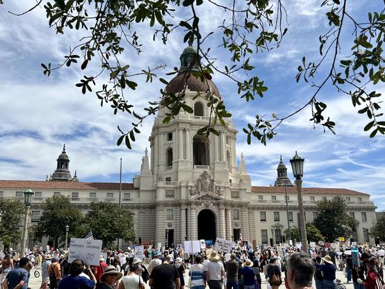 At NoKings protest, in front of Pasadena City Hall with Judy Chu speaking. The beautiful Mediterranean-style City Hall building is flanked by wispy high clouds in a blue sky