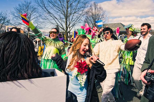 Woman with long bland hair sings on (wireless?) micropone surrounded by more paradge goers including a man in white holding an American football in an outstretched hand