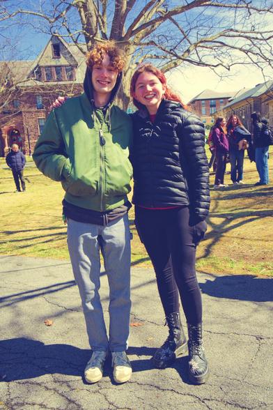 A man with fuzzy hair and green jacket and grey pants on the left with a a woman wearing dark leggings and a dark puffer jacket on the right