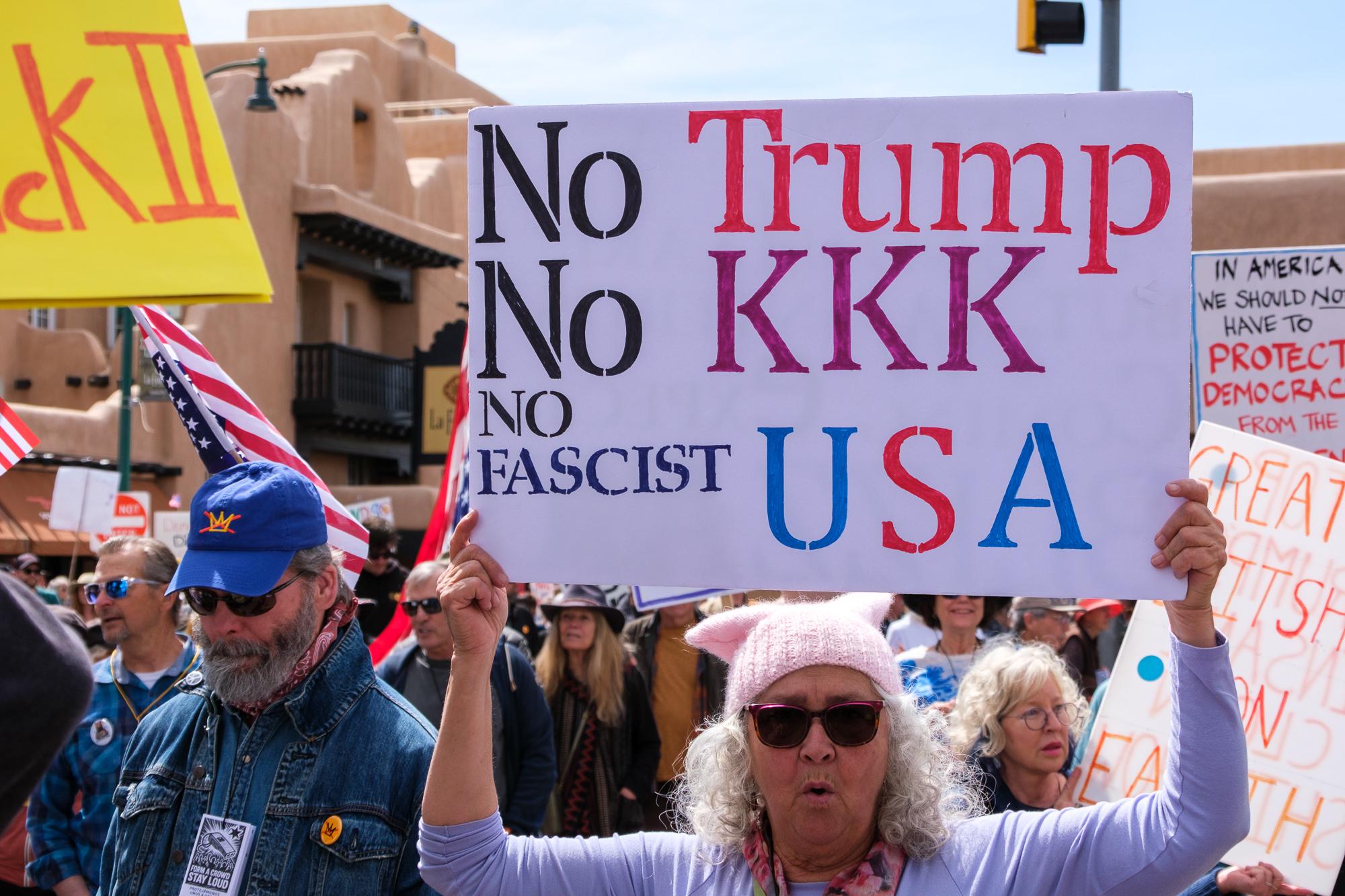 Woman in a pussy hat holding a sign that says “No Trump, No KKK, No Fascist USA.  She is in the foreground with many other marchers all around her.