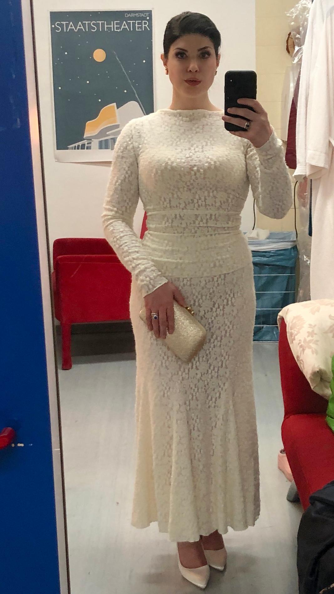Selfie mirror-portrait of a woman with dark hair styled into a chic updo, with red lipstick, and false eyelashes, wearing a fluffy white lace top and skirt combo with a boat neckline and long sleeves. In her left hand she is holding a smartphone, in her right a cream-white clutch purse. Megan Marie Hart in her dressing room at the Staatstheater Darmstadt after the premiere of Felix Weingartner’s opera “Kain und Abel” on March 28, 2026. CC BY-SA 4.0