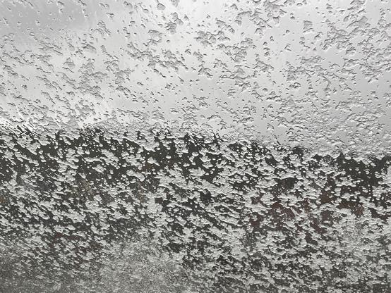 Ein von innen fotografiertes Dachfenster voller Hagelkörner, schemenhaft ist die Stadt zu sehen.