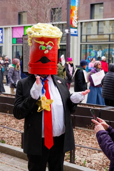 Fake Donald Trump with his head made of an orange-painted sheetrock bucket with a face pointed on and a comically ill fitting suit and long red tie stands in front of empty planter while a woman holds up a cell phone and a crowd flows on the other side of the ithaca commons