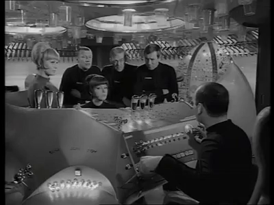 Szene aus der Serie "Raumpatrouille Orion", schwarz-weiß. Die Besatzung steht bzw. sitzt um eine blinkende Maschine herum. 