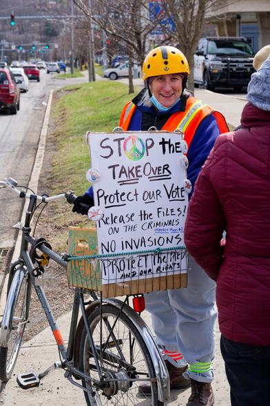 Woman with bright yellow helmet and safety vest on top of a blue jacket is standing behind a bike and i think a woman in a purple coat and holds a sign that says STOP THE TAKEOVER Protect or Votes Release the files on the Criminals NO INVASIONS