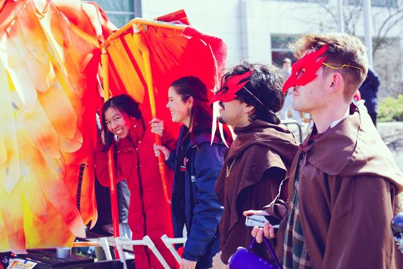 On the far left a mass of pipes and feathers held up by a Chinese woman who is looking at me with a big smile and to her right a white woman wearing a blue jacket and two white men wearing orange phoenix masks and brown cloaks.