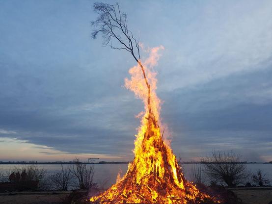 Photograph taken by my sister of an Easter bonfire. It is shaped like a witch's hat. A dusky sky and river with a few bushes at the shore can be seen in the background.