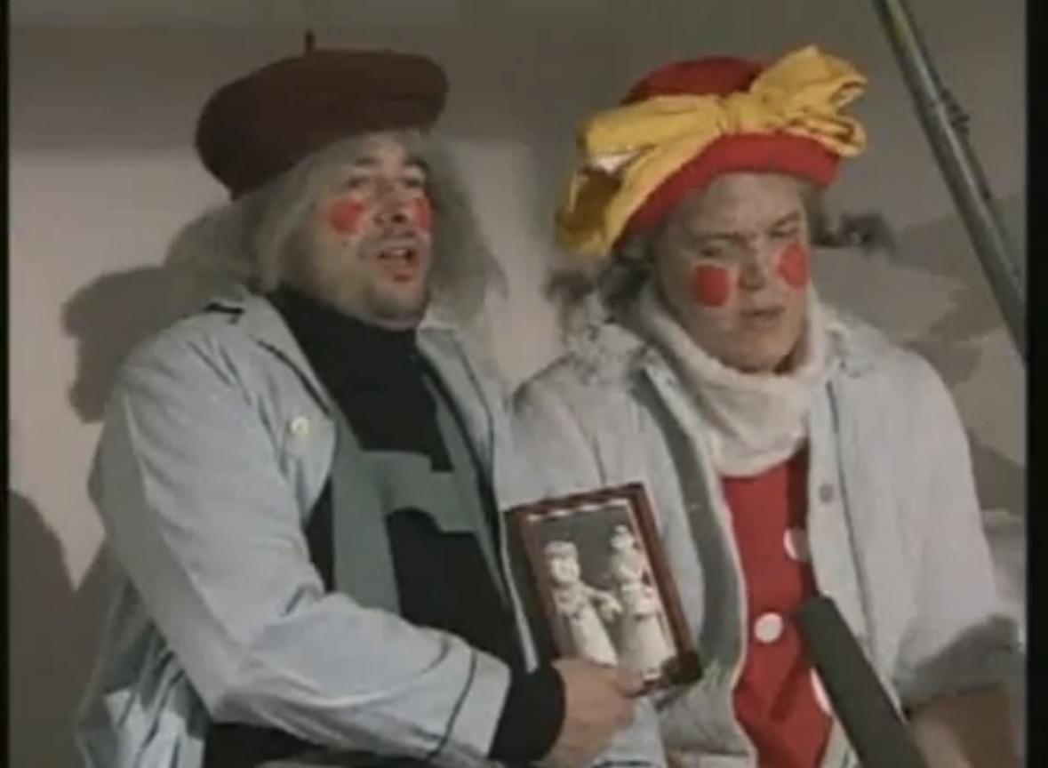 A low quality frame from a tv show. Two people in a hospital setting, both wearing red circles on their cheeks and otherwise covered in circular markings. They are sitting on a hospital bed wearing white gowns. Tipp holds a framed black and white photograph of two dolls. There’s a microphone in the frame.