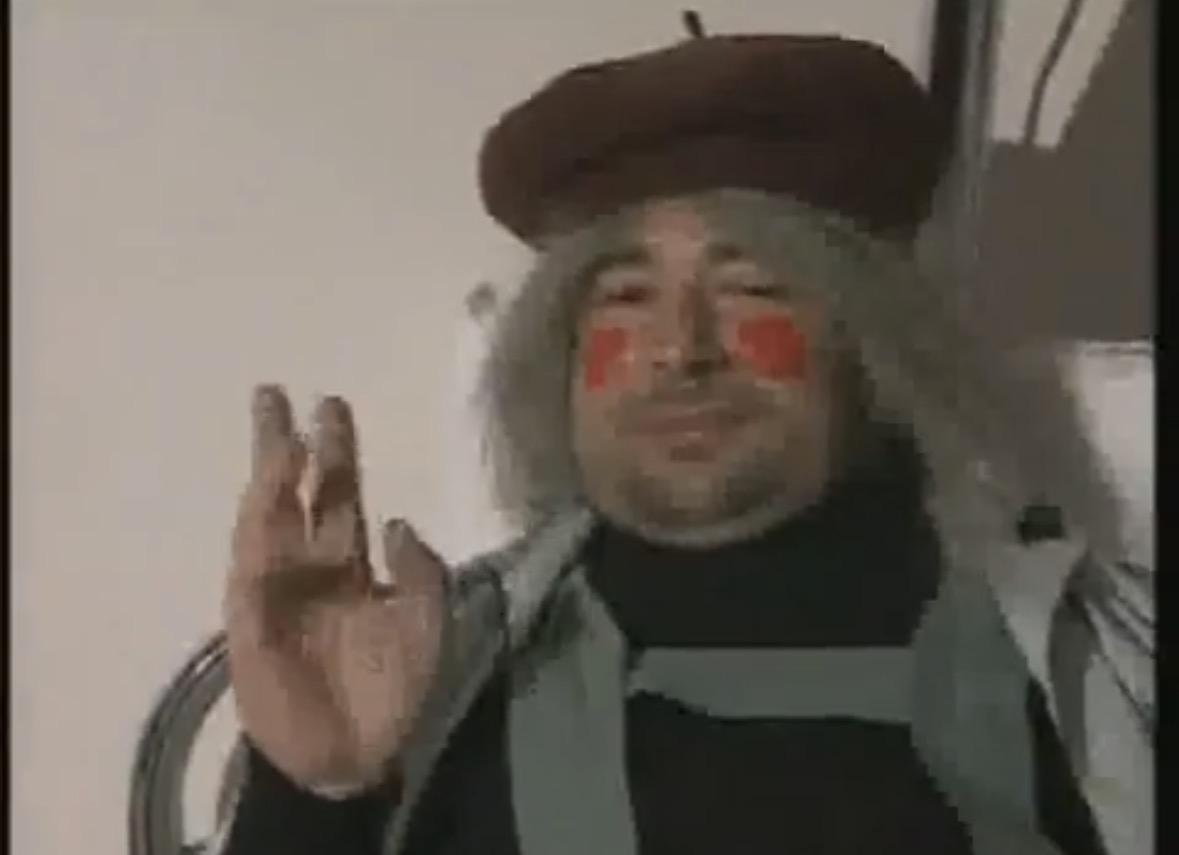 A middle aged man with a stubble, holding up his right hand. Has red circles painted on his cheeks. Low quality frame from video.