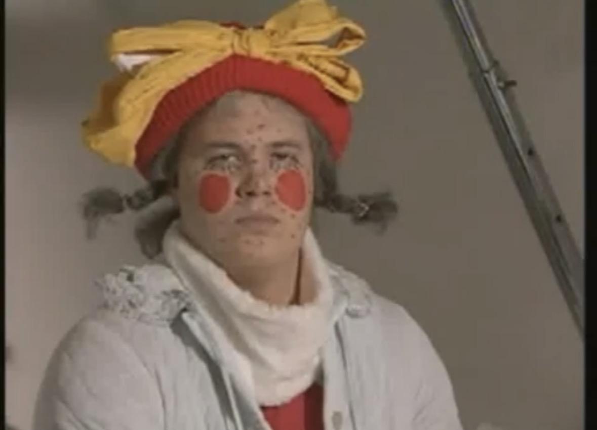 A middle aged man dressed in hospital gown and wearing pigtails and a feminine headwear. His face is full of black dots and he has two red circles on his cheeks.