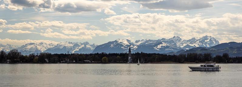 
vorne: die Wasserfläche eines großen Sees mit einem Boot. Das andere Ufer und im Hintergrund eine Kette hoher Berge mit schneebedeckten Wipfeln.