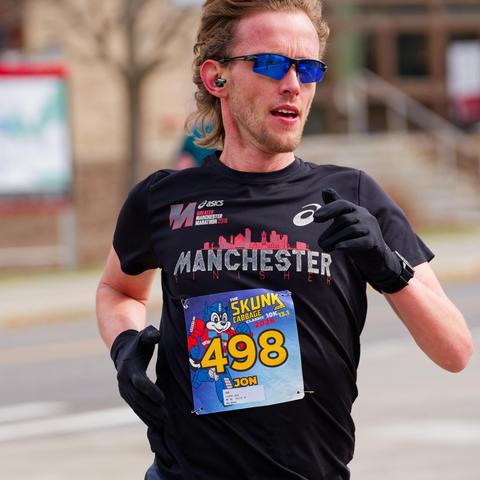 Close shot of runner with black shirt that says MANCHESTER with a red skyline above it, bib number 498, short light colored hair and black gloves with a beige building with a map sign next to it.
