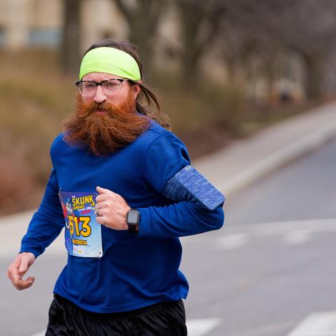 Man with thick reddish beared and blue shirt, bib number 313, green bandanna and dark pants with a slope of trees and bushes behind him