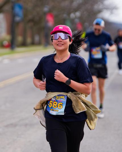 Woman with pink hat, mirror shades, dark shirt, dark pants, bib number 586 and light colored garmet around her waist running on road with two men in the background as well as blue and red banners and leafless trees