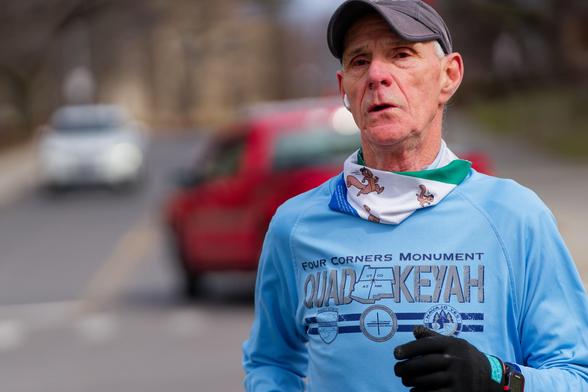 Older man with billed cap and scarf and blue shirt with one visible gloved hand and cars moving in the background