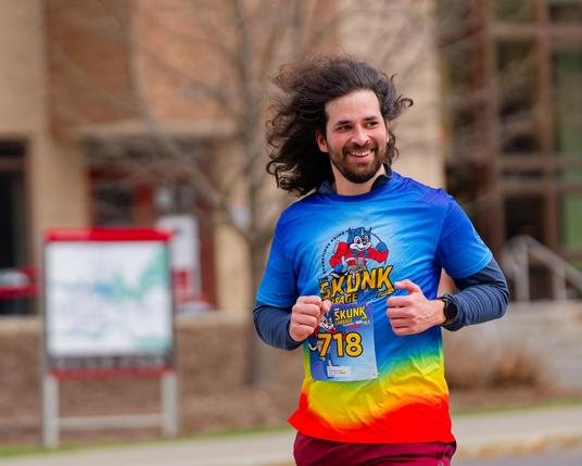 Man with long hair, beard and big smile with the rainbow colored run shirt for this year seen from the hips up with a building with a map sign out front behind him