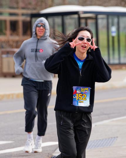 Woman with dark shades and dark clothes makes double v-sign with another runner with black pants and light grey hoodie behind her and a bus stop