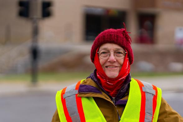 Woman with red wool cap and high viz vest over brown jacket with beige building and road and background