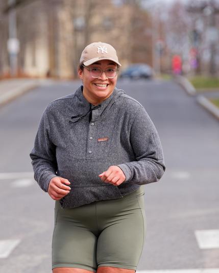 Woman with big smile, billed cap, mid-grey jacket and shorts with tranquil campus background