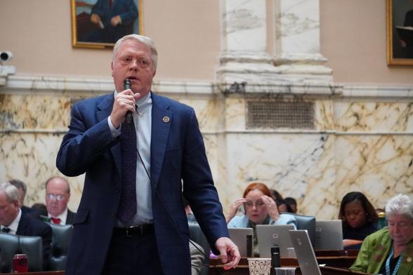 Man with short white hair and dark blue suit with darker blue tie and light blue shitrt holds microphone in one hand with more people sitting with computers behind him.  Behind them all is a wall where the lower part and columns are made of marble with framed portraits hanging on a cream colored section above.