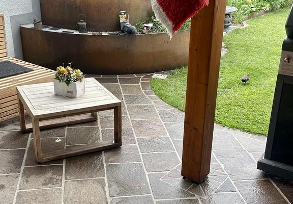 Gefliester Gartensitzplatz mit einem quadratischen Tischchen aus Holz. Darunter hat die Amsel einen weissen Haufen gesetzt. Sie selbst rennt auf den Rasen davon.