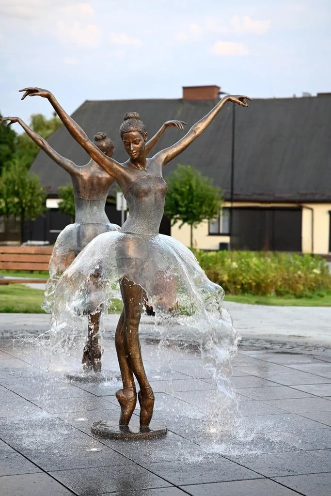 A photograph of a bronze fountain sculpture featuring two ballerinas in a dance pose. The water from the fountain is engineered to spray out from their waists, creating the visual effect of translucent, shimmering tutus. The statues are set on a gray tiled plaza with trees and a building in the background.