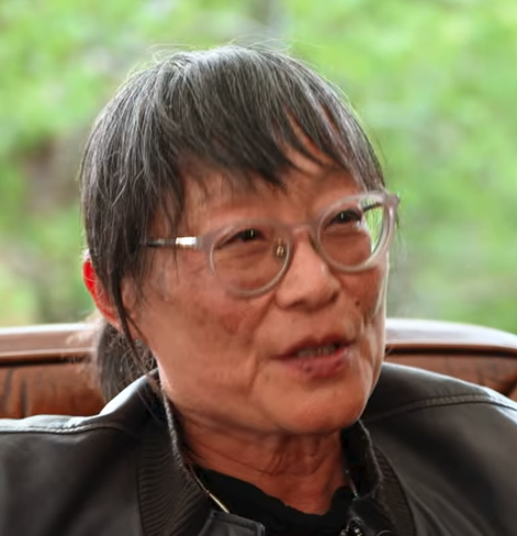 Headshot of asian woman with medium length slightly greyed dark hair tied back in the back with round plastic glasses and leather jacket and dark shirt
