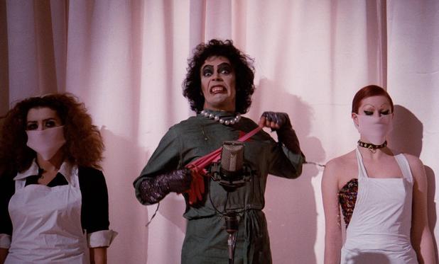Picture of Tim Curry as Frank N Furter from Rocky horror picture show in surgical gown glittery gauntlets pearls and snapping rubber gloves while standing in front of a microphone flanked on either side by Columbia and Magenta