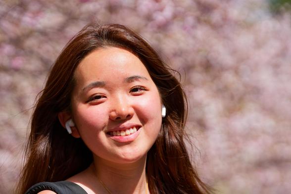 Smiling asian woman wearing airpods with brown hair with very blurry pink tree flowers behind her.