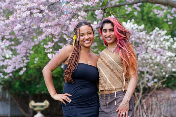 Two women with slightly dark skink pose together:  the one on the left has a dark blue dress with bare shoulders and i think dreadlocks and the one on the right has long hair colored red at the top gradienting to brown with a brown shirt with diagonal stripes and a gorgeous pink sakura tree is behind them.