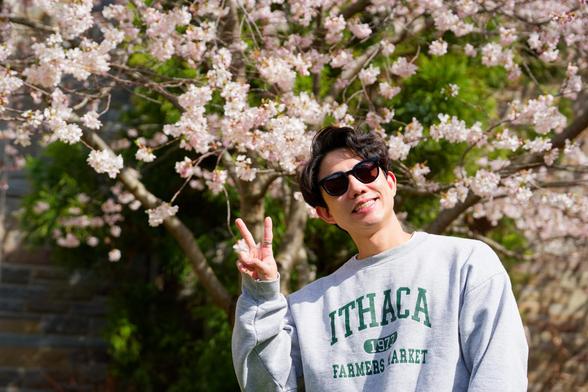 Asian man with moderate length hair and dark glasses has farmer's market shirt and makes V sign under pink flowers