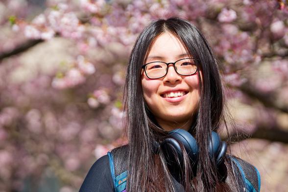 Asian woman with black-framed glasses wears headphones under long hair with lovely pink petals behind her