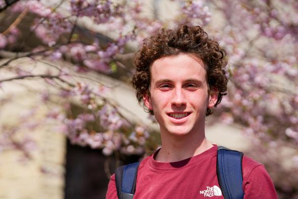 man with moderate curly brown hair, smile, red shirt and backpack straps with a light dusting of pink flowers behind and the blurred out shapes of a stone building with large and small windows